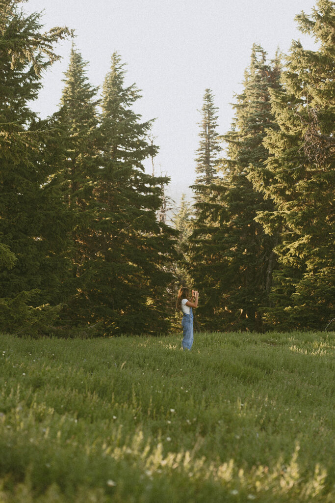 A woman is standing in a grassy field surrounded by trees.