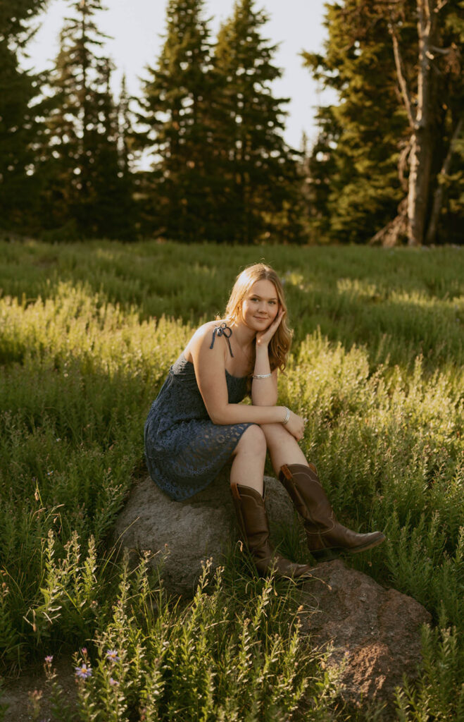 A woman in a dress and cowboy boots is sitting on a rock in a field.