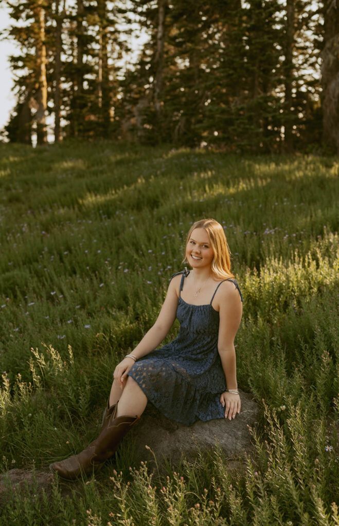 A woman in a dress and cowboy boots is sitting on a rock in a field.