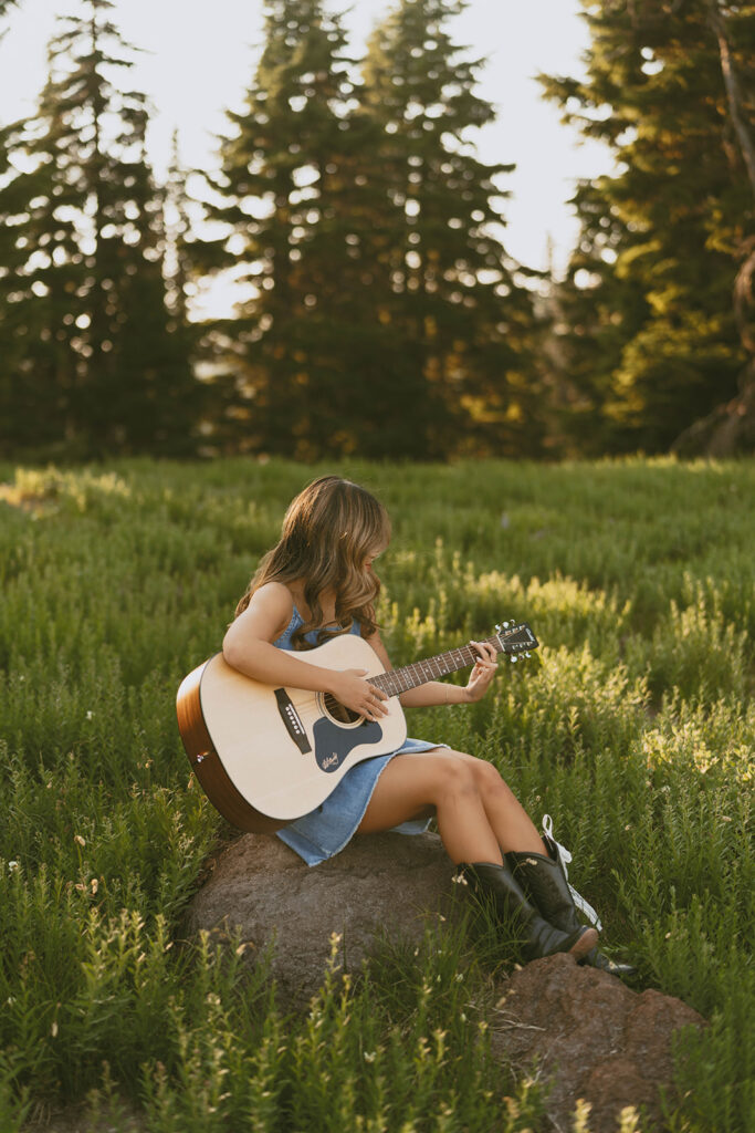 A woman is sitting on a rock in a field playing a guitar.