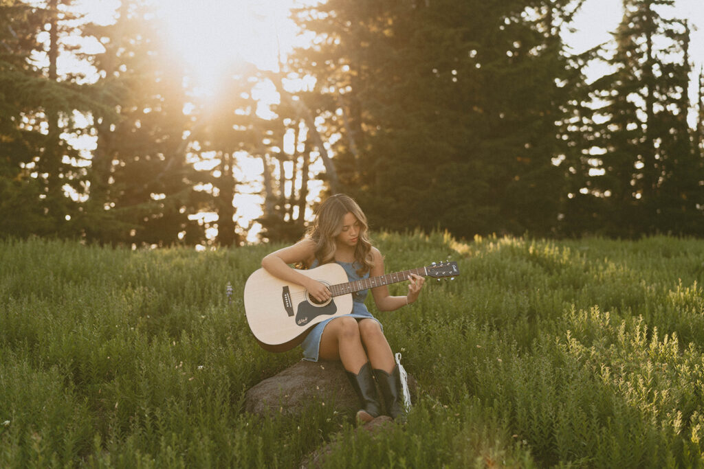 A woman is sitting on a rock in a field playing a guitar.