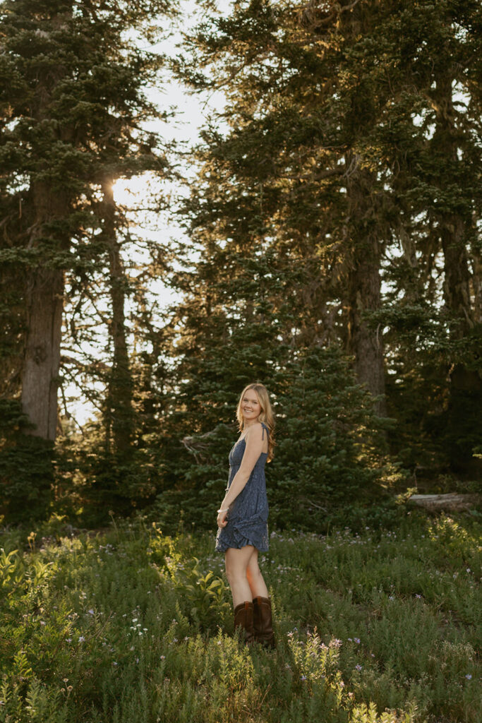 A woman in a blue dress and brown boots is standing in a field in the woods.