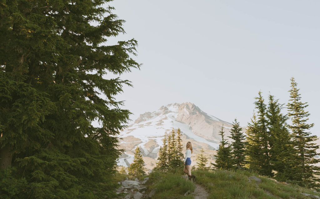 A woman is standing on top of a grassy hill looking at a mountain.