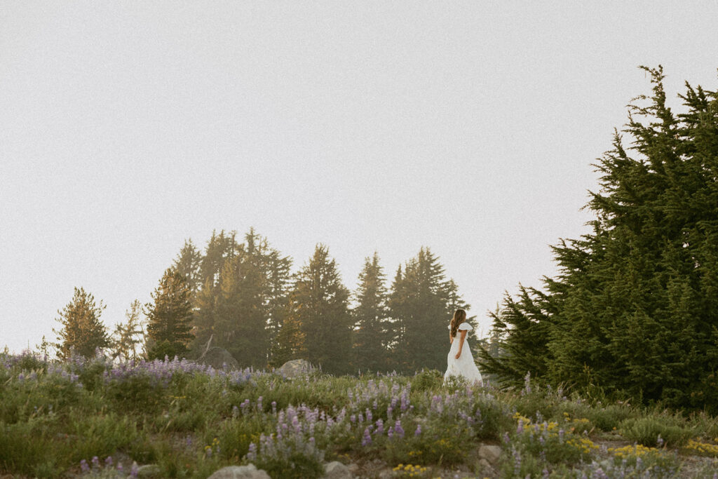 A woman in a white dress is standing in a field with trees in the background.
