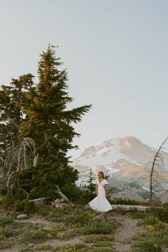A senior in a white dress is standing in front of a mountain.