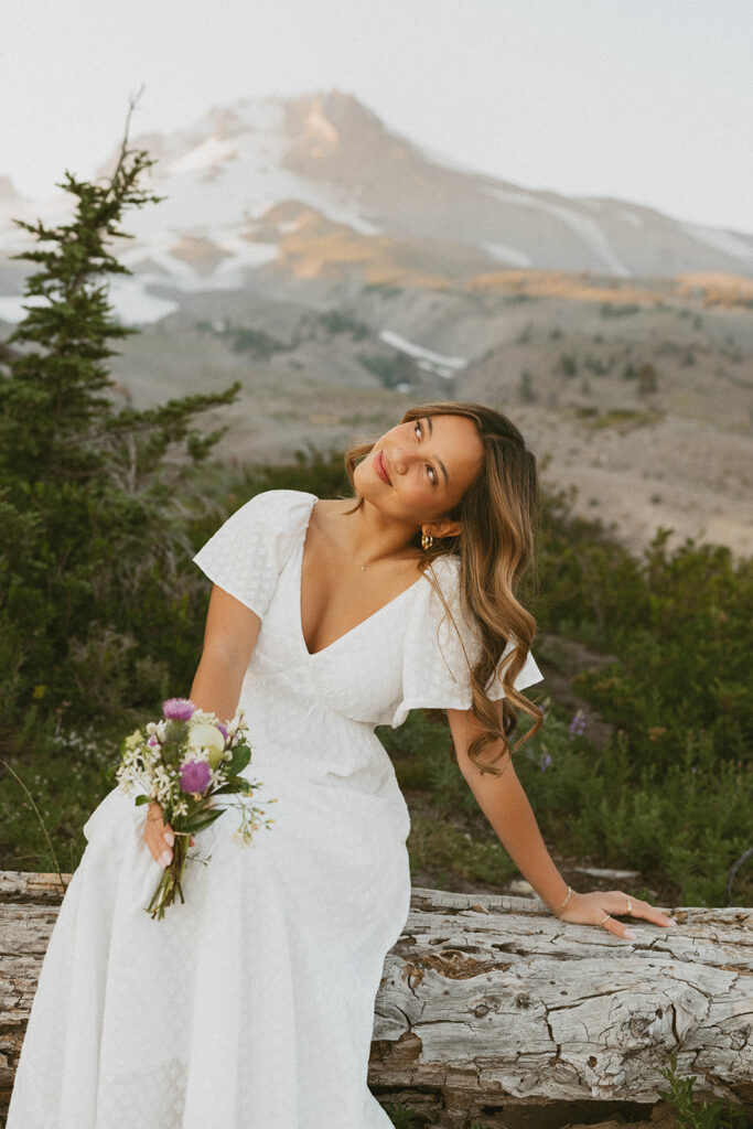 A woman in a white dress is sitting on a log holding a bouquet of flowers.