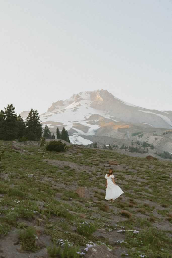 A woman in a white dress is standing in a field with a mountain in the background.