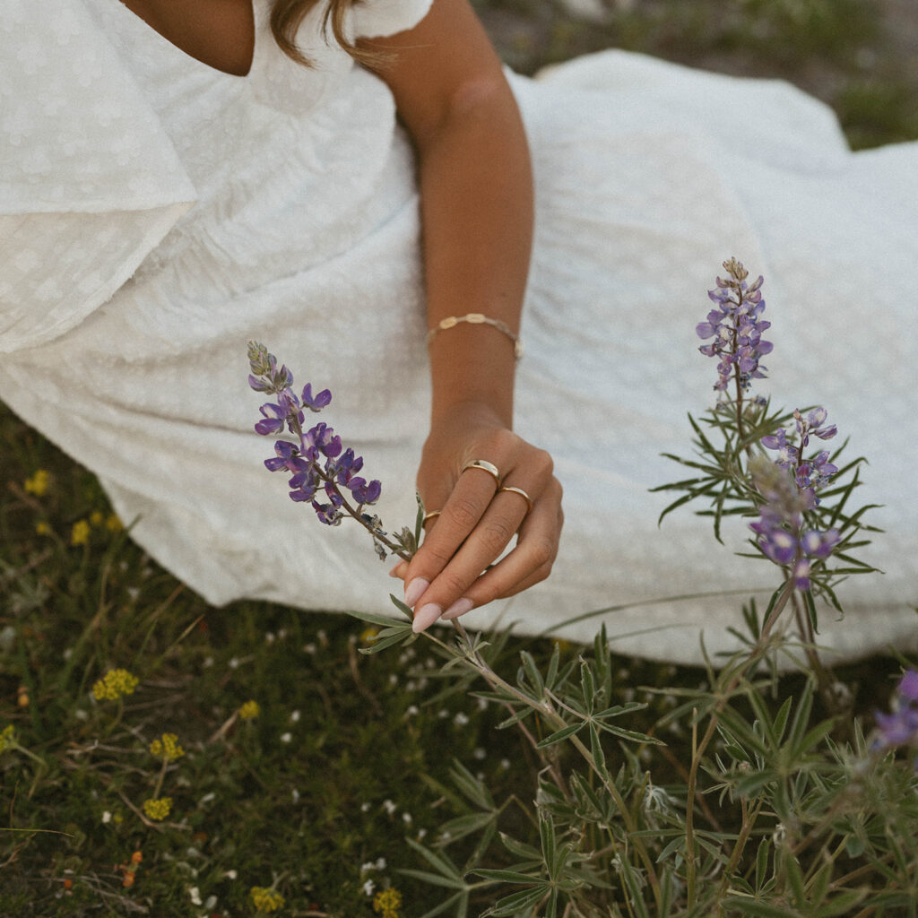 A woman in a white dress is laying on the ground picking purple flowers.