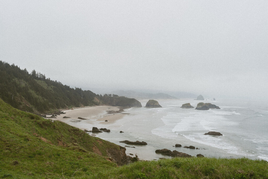 Coastal landscape with sea stacks, sandy beach, and forested cliffs under a cloudy sky