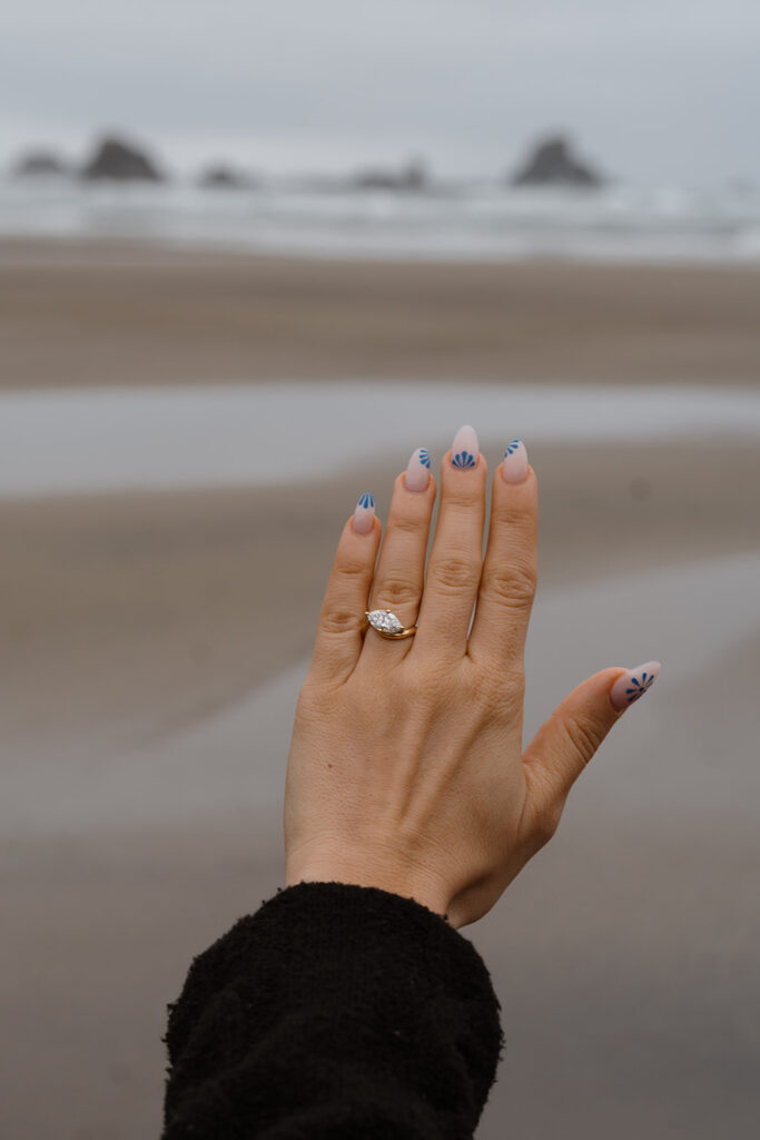 Hand with engagement ring extended toward ocean and sea stacks