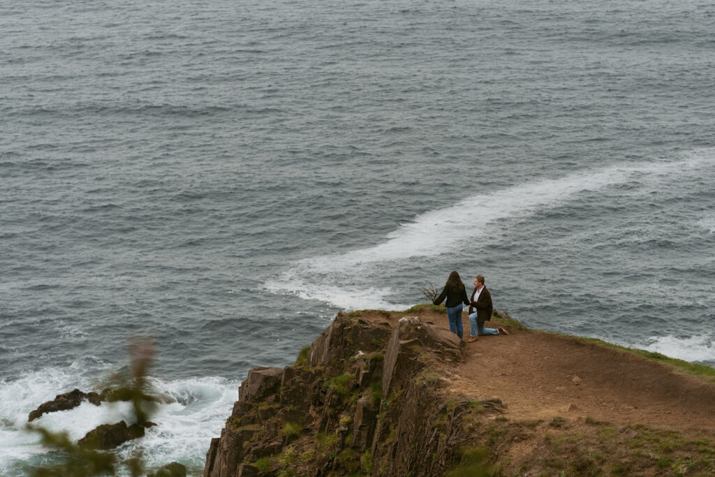 Couple at the edge of a rocky cliff above the ocean for a beach proposal
