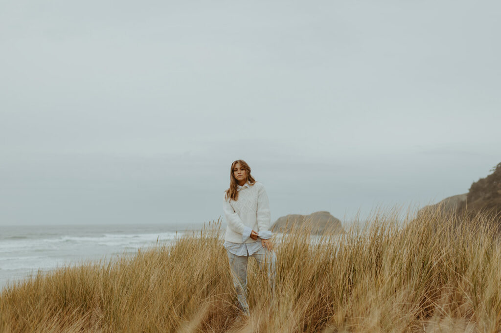 Young woman in white sweater stands in golden coastal grass in coastal Portland
