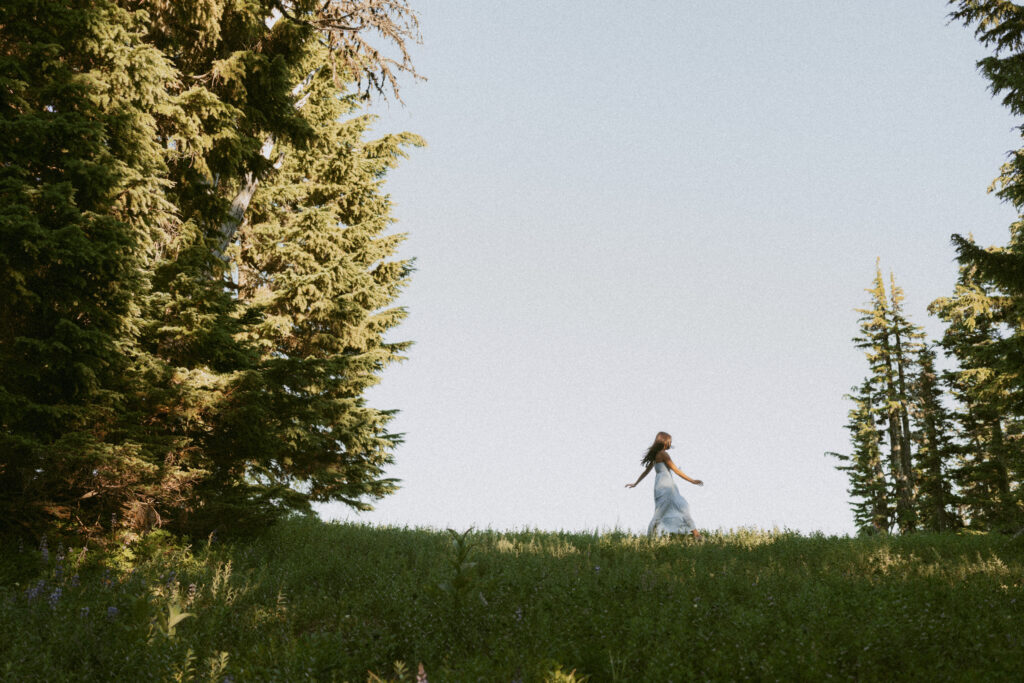 Minimalist Mount Hood senior portrait of girl in flowing white dress walking across open meadow against cloudy sky backdrop