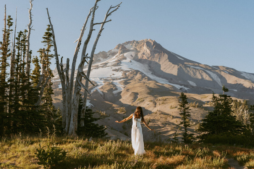 girl in white maxi dress with arms outstretched against Mount Hood mountain vista and dead trees for dramatic outdoor portrait