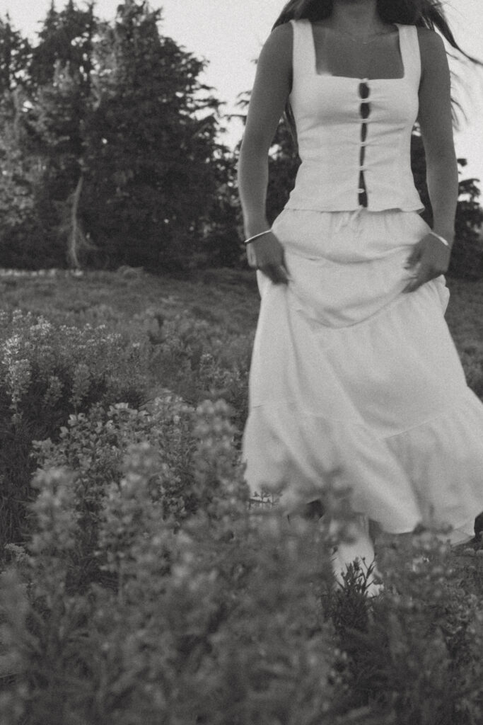 artistic black and white senior portrait of girl in white dress walking away through Mount Hood wildflower field