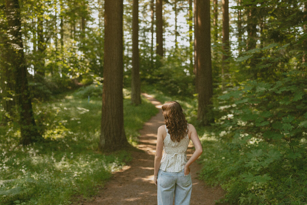 girl in white lace top and jeans walking down forest trail through tall trees during Portland senior portrait session at Hoyt Arboretum