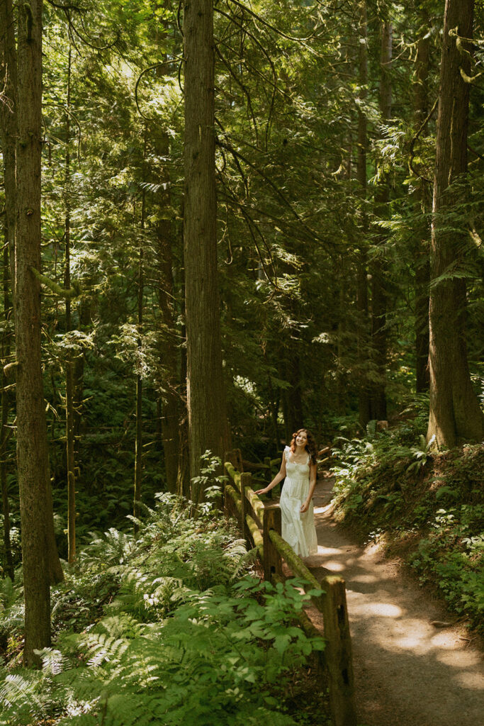 Hoyt Arboretum Portland senior photos featuring girl in white dress walking and smiling along soft dirt path through towering redwood forest with dappled sunlight