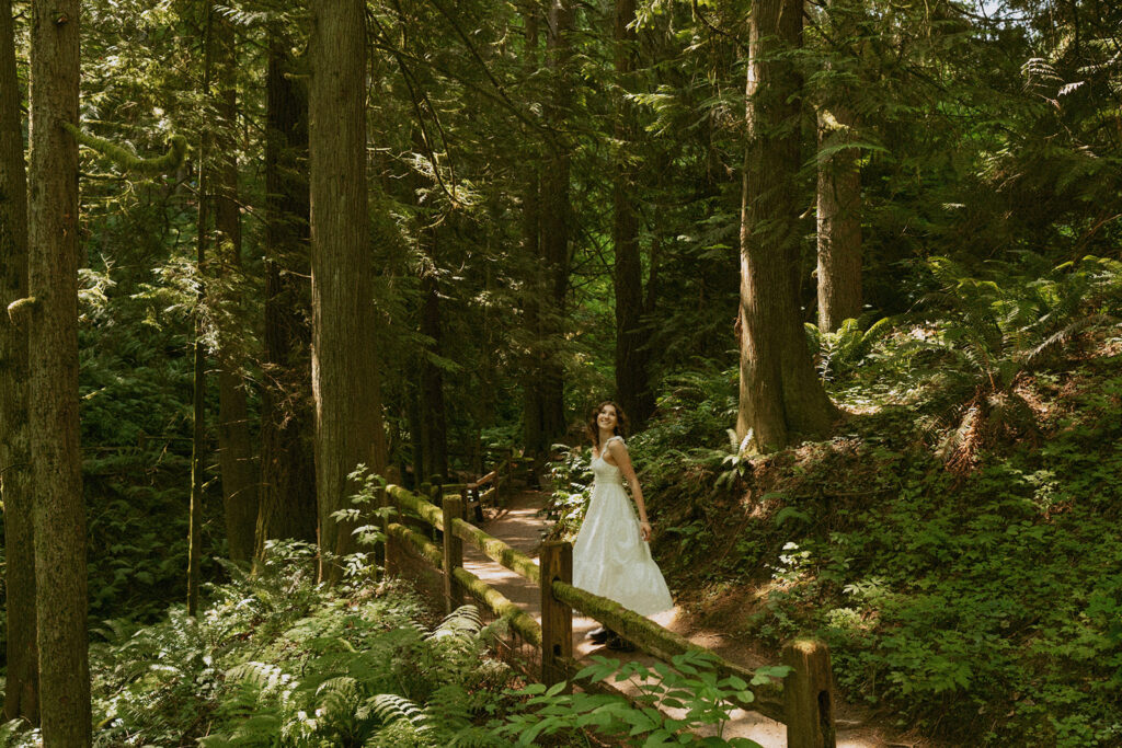 Hoyt Arboretum Portland senior photos featuring girl in white dress crossing wooden bridge through towering redwood forest with dappled sunlight