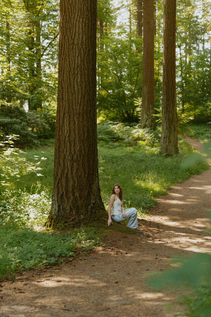 Portland senior photographer captures girl in white polka dot top and blue jeans sitting against massive tree trunk at Hoyt Arboretum with natural lighting
