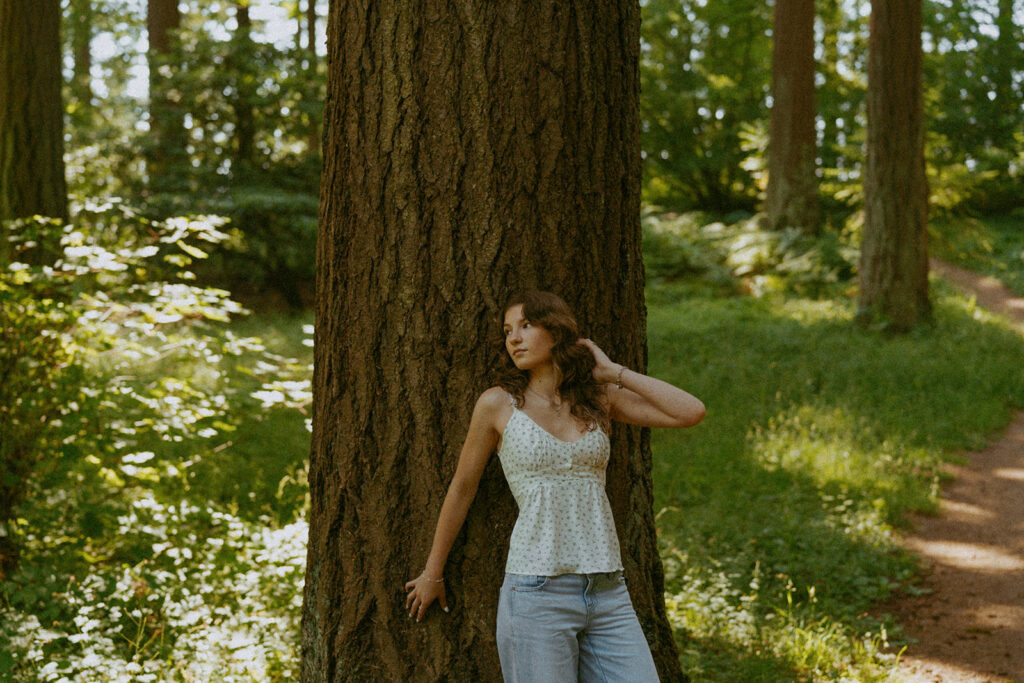 Portland senior photographer captures girl in white polka dot top leaning against massive tree trunk at Hoyt Arboretum with natural lighting