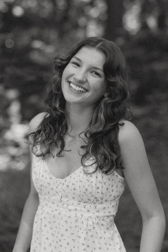 Black and white artistic senior portrait of smiling girl in white dress among towering trees at Hoyt Arboretum Portland