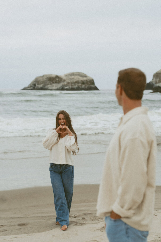 Jentrie makes a heart shape with her hands while Brayden looks at her on the beach