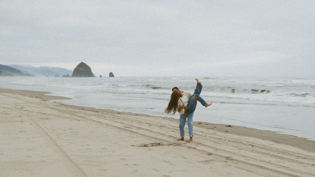 Man carrying woman while standing on the beach with ocean waves behind.