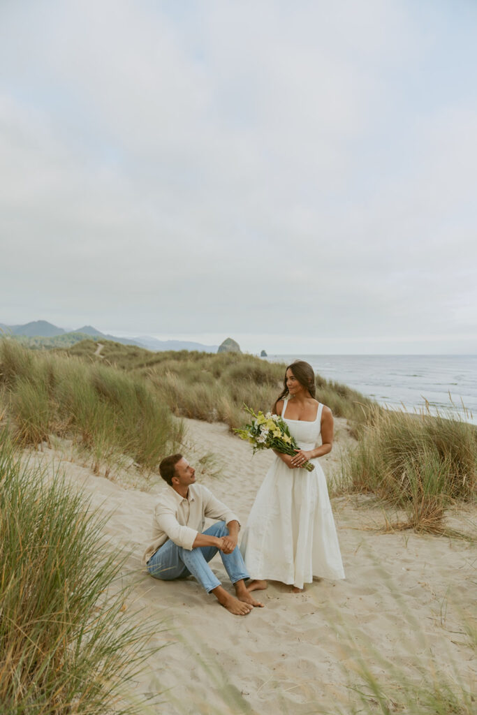 Woman in white dress holding flowers while man sits on the sand dunes