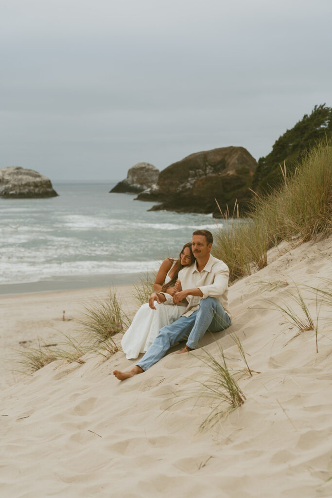Couple embraces and laughs while sitting down on the shore, with rocky cliffs visible behind them