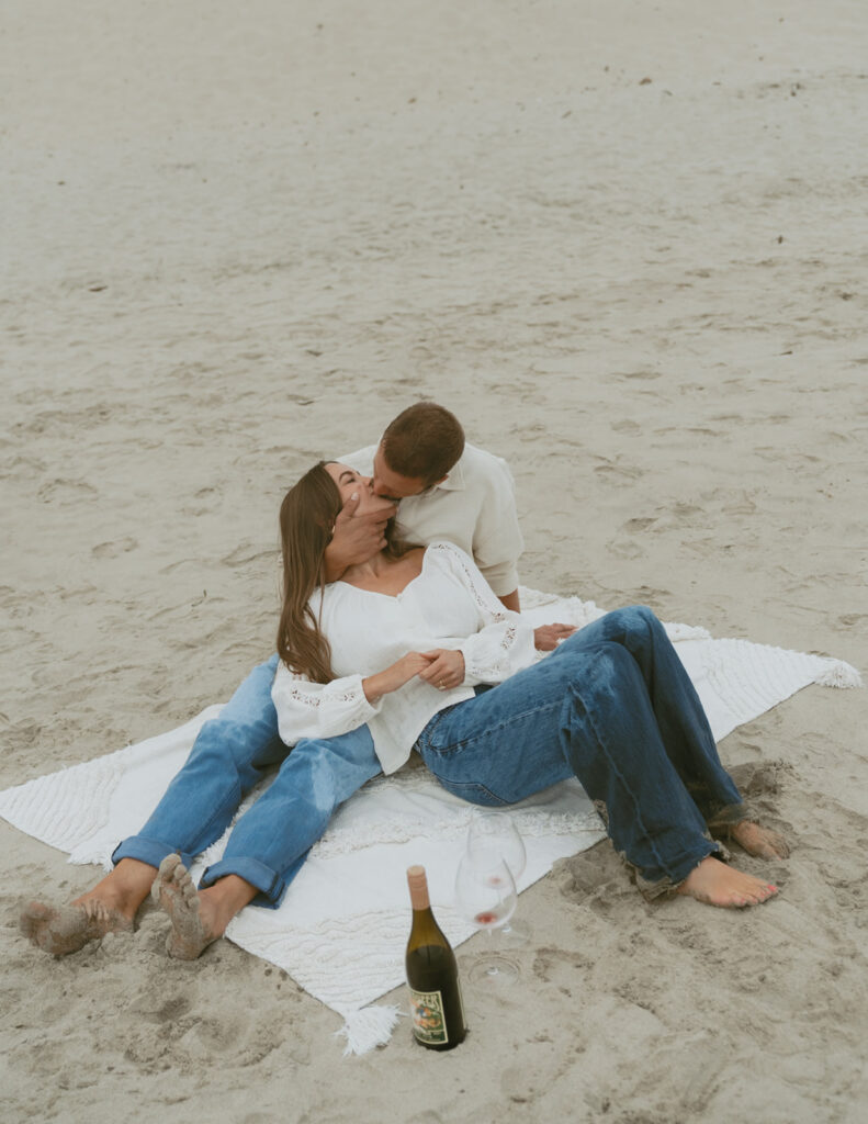 Couple kissing on a blanket at the beach with a wine bottle nearby
