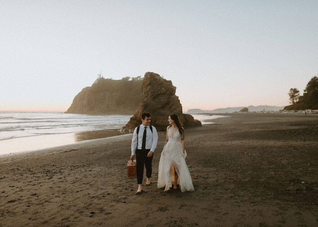 Bride and groom walking barefoot on beach with picnic basket