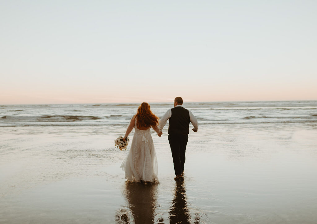 Bride and groom walking hand in hand into the ocean at sunset