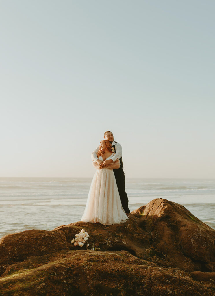 Groom embracing bride on seaside rocks during Oregon elopement