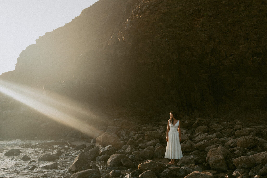 Woman in white dress standing on rocky beach with sunlight rays
