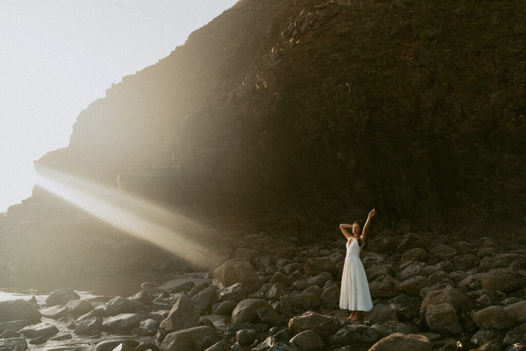 Woman in white dress standing on rocky beach at sunrise