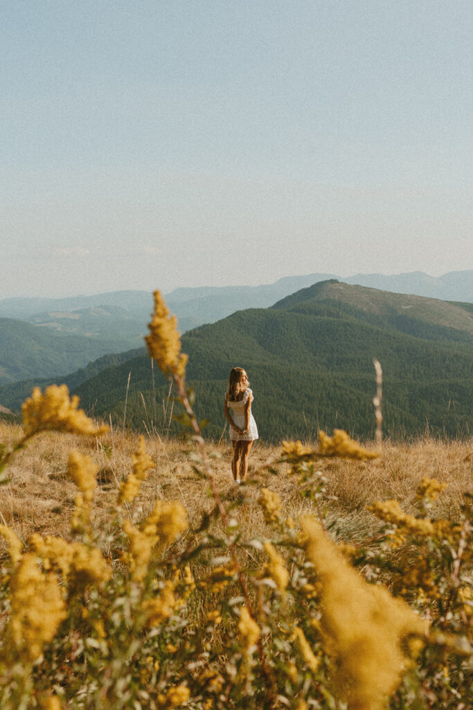 A senior girl stands in a golden field overlooking mountains, captured by a Salem senior photographer