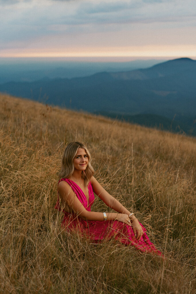 A senior smiles while sitting in tall grass with mountain views