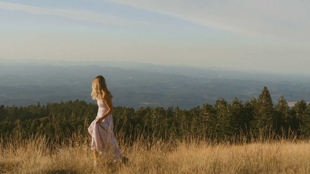 A senior girl walks through tall grass on a mountain ridge