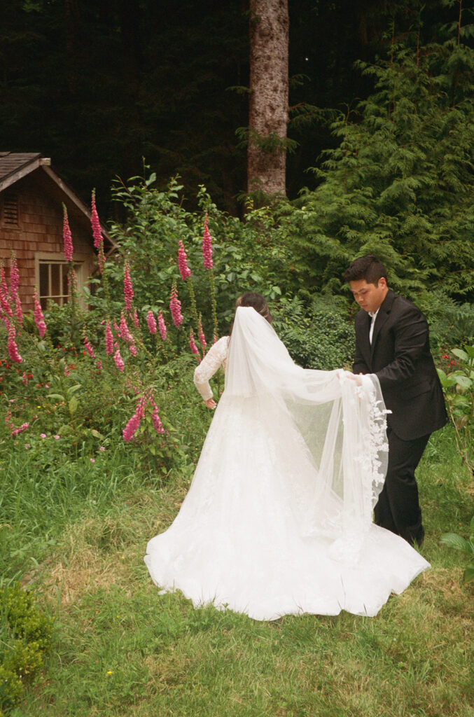 Groom holding the bride’s veil in a garden by a cottage