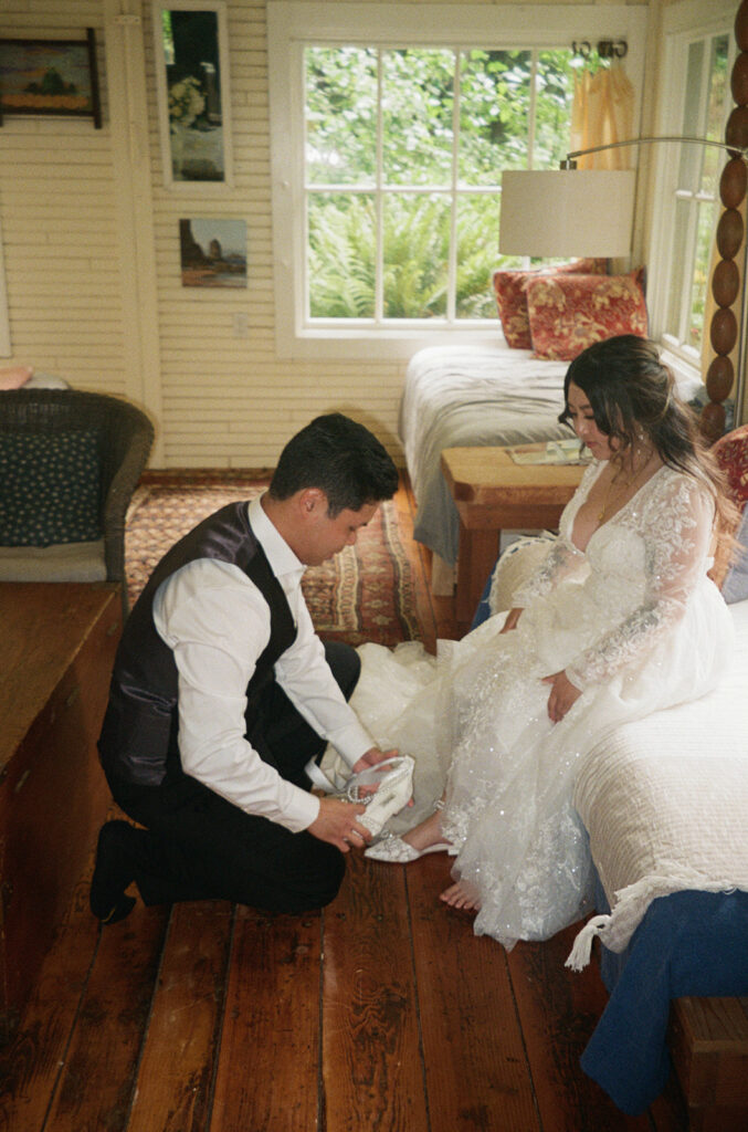 Groom helps bride put on her shoes in a cozy cabin bedroom