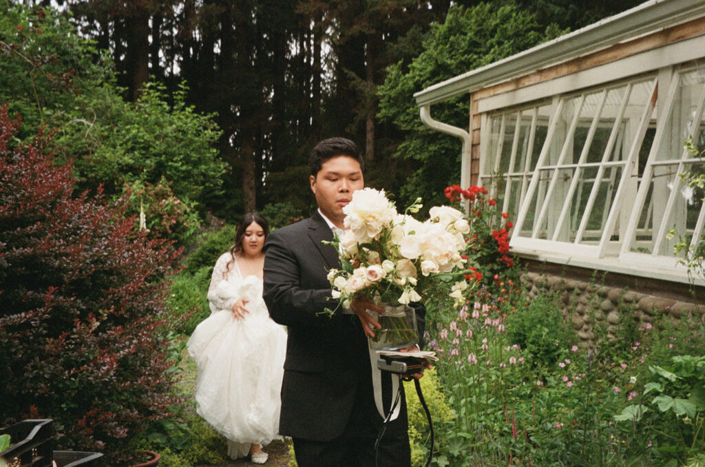 Groom carrying a large bouquet with bride walking behind in a garden