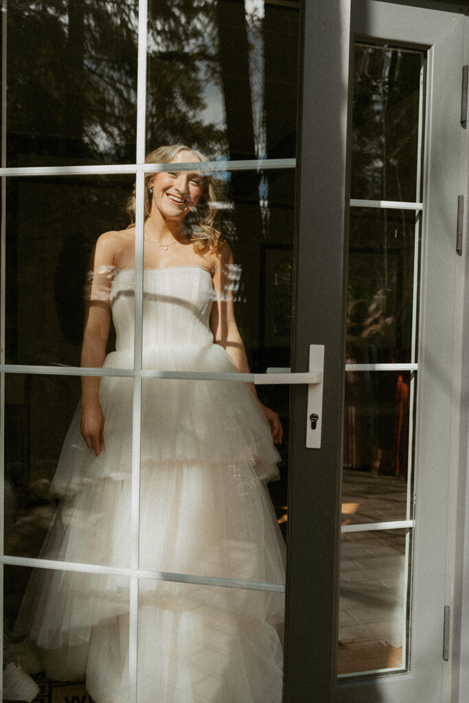 Bride in layered gown smiling through glass door