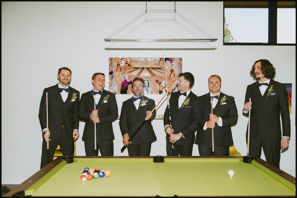 Groomsmen in black tuxedos posing with pool cues around a pool table before the wedding