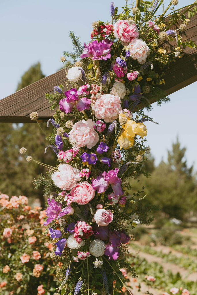 Colorful floral installation on a wooden ceremony arch, featuring pink peonies, purple iris, and yellow blooms