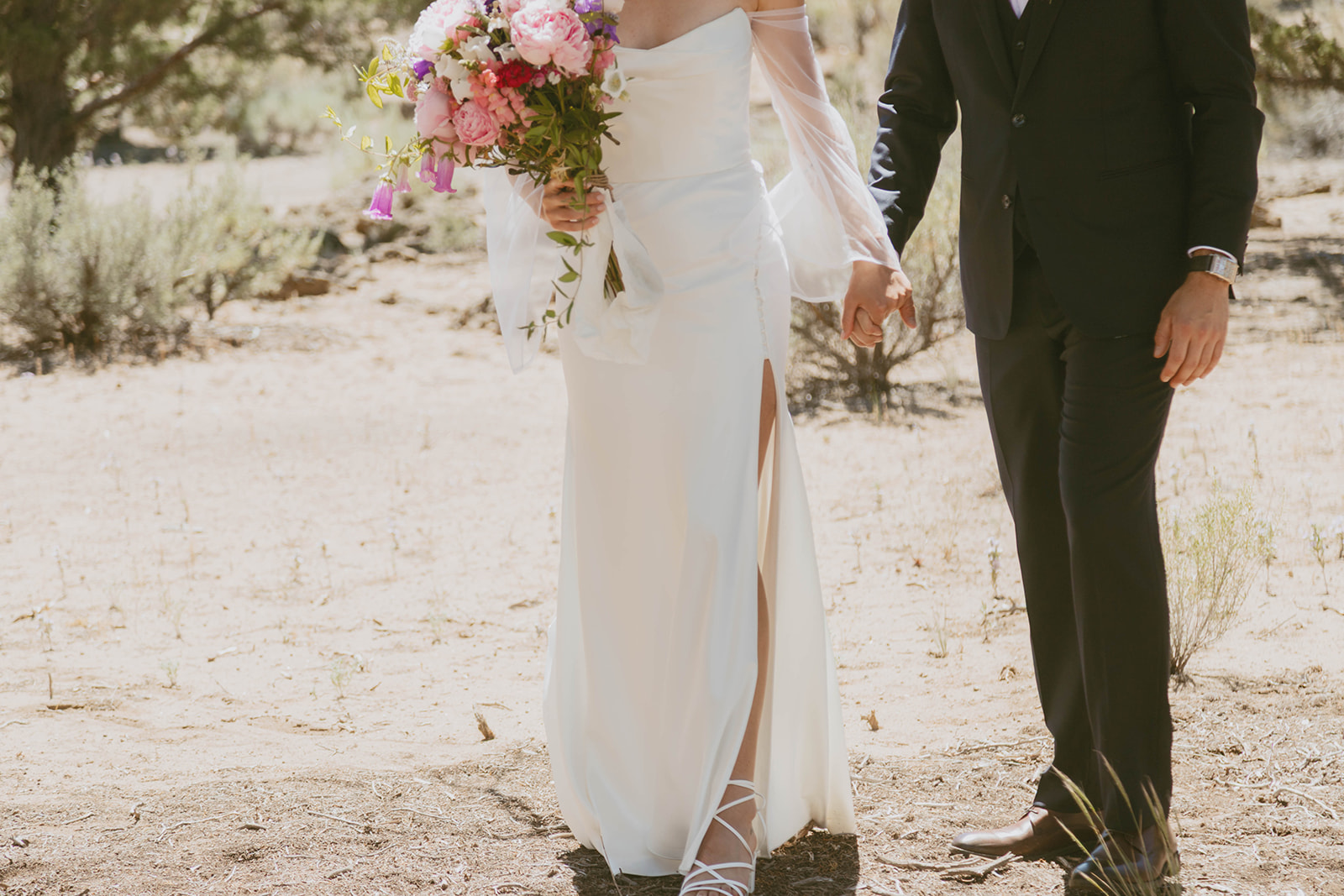 Bride and groom holding hands in the desert, bride's high-slit wedding dress and bouquet of pink and purple flowers in focus