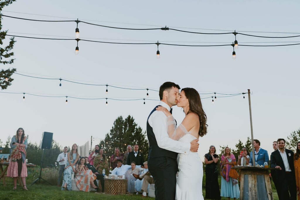 Bride and groom sharing a kiss during their first dance at golden hour, wedding guests watching in the background