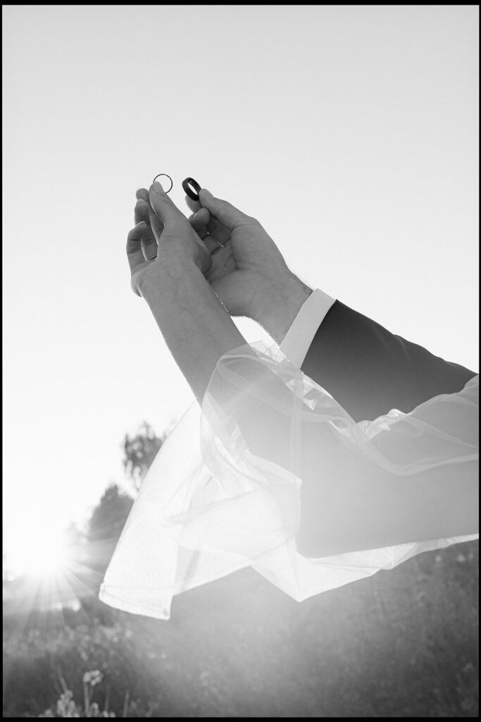 Black and white film photo of bride and groom's hands holding wedding rings against a sunlit sky, veil blowing in the wind
