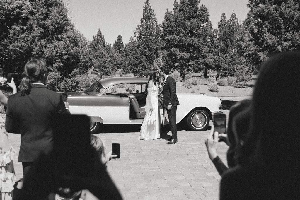 Black and white film photo of guests photographing bride and groom kissing beside a vintage car after their wedding ceremony