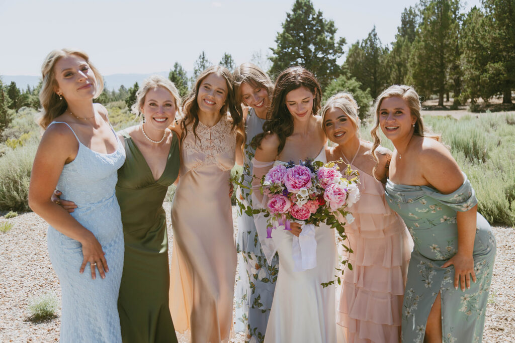 Bride surrounded by her bridesmaids in mismatched dresses, holding a bouquet of pink and white peonies